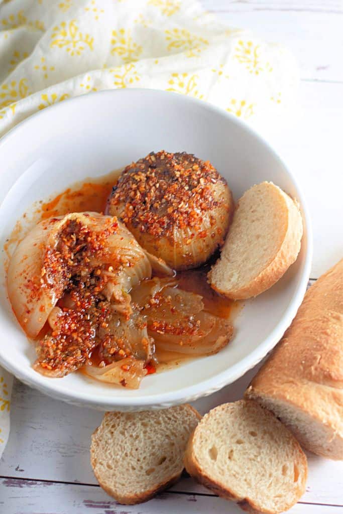 A white bowl contains two whole roasted onions with a golden-brown, caramelized surface, topped with a seasoning mix of crushed red pepper, garlic, and spices.