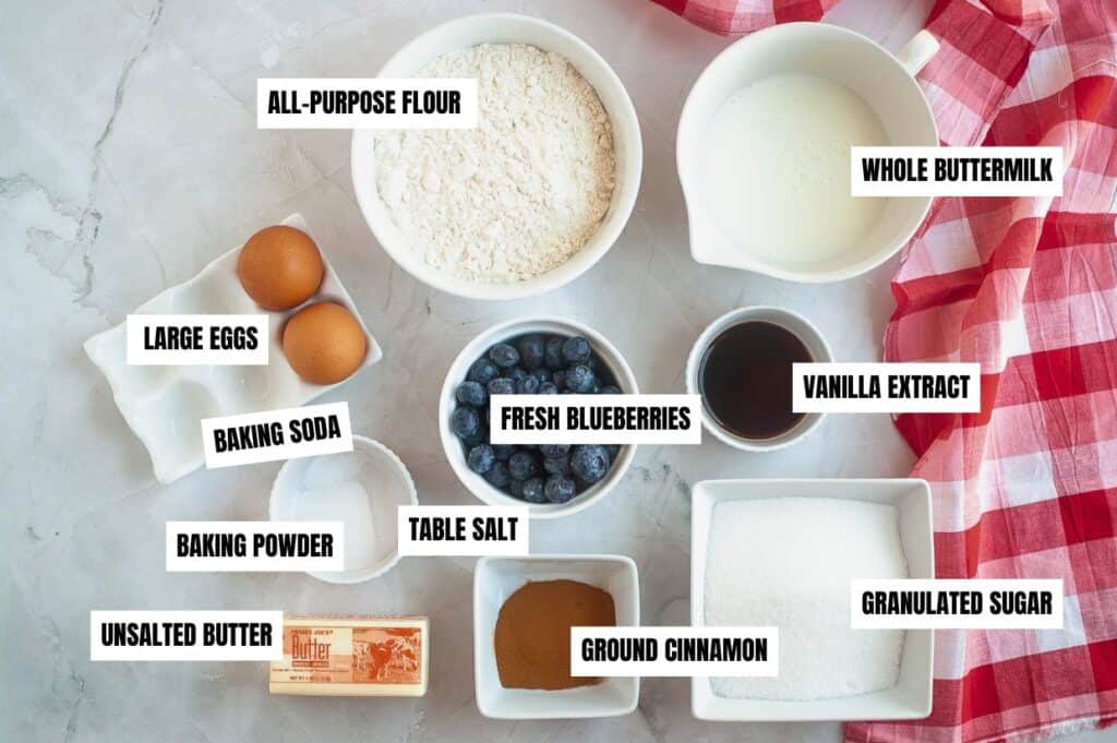 A flat-lay image of ingredients for Blueberry Boy Bait, including all-purpose flour, whole buttermilk, fresh blueberries, granulated sugar, large eggs, unsalted butter, vanilla extract, ground cinnamon, baking powder, baking soda, and table salt, arranged on a marble surface with a red and white checkered cloth.