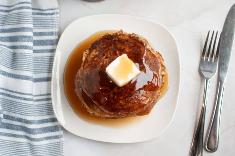 A stack of golden-brown buttermilk oatmeal pancakes on a white plate, topped with a pat of melting butter and drizzled with maple syrup.