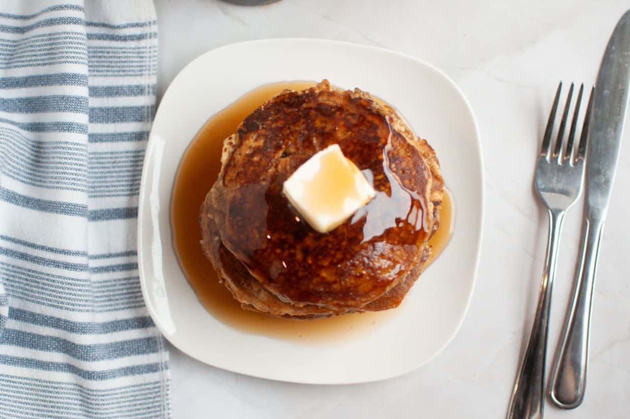 A stack of golden-brown buttermilk oatmeal pancakes on a white plate, topped with a pat of melting butter and drizzled with maple syrup.