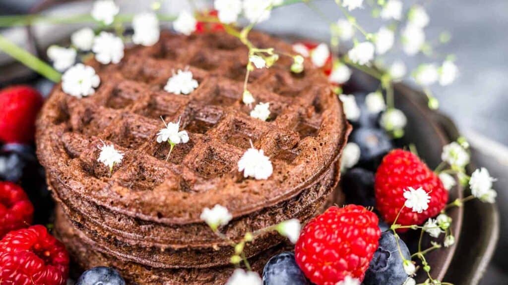 Stack of chocolate chaffles topped with flowers, served with fresh raspberries and blueberries.
