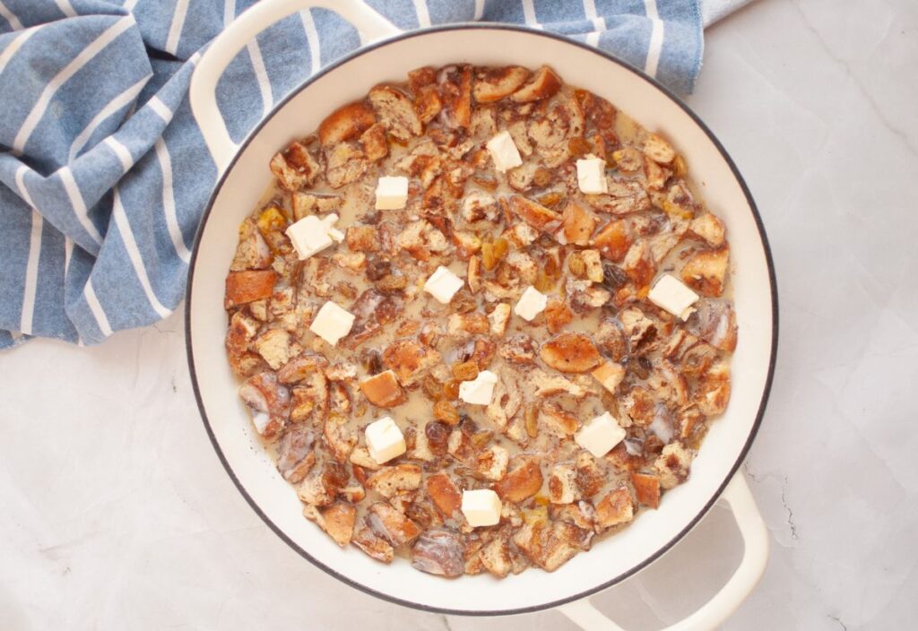 A baking dish layered with bakery cinnamon rolls and golden raisins with added custard base and cubed butter scattered on top.