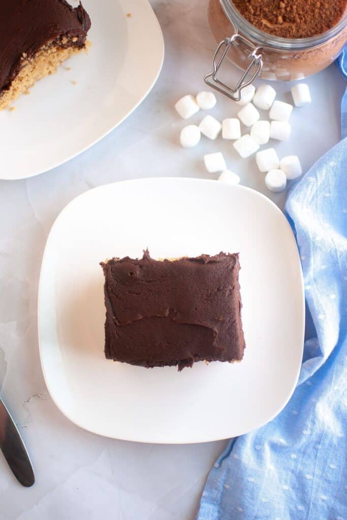 A slice of Graham Cracker Cake with rich chocolate frosting sits on a white plate. In the background, mini marshmallows, a jar of cocoa powder, and a partially sliced cake are arranged on a marble surface with a blue cloth.