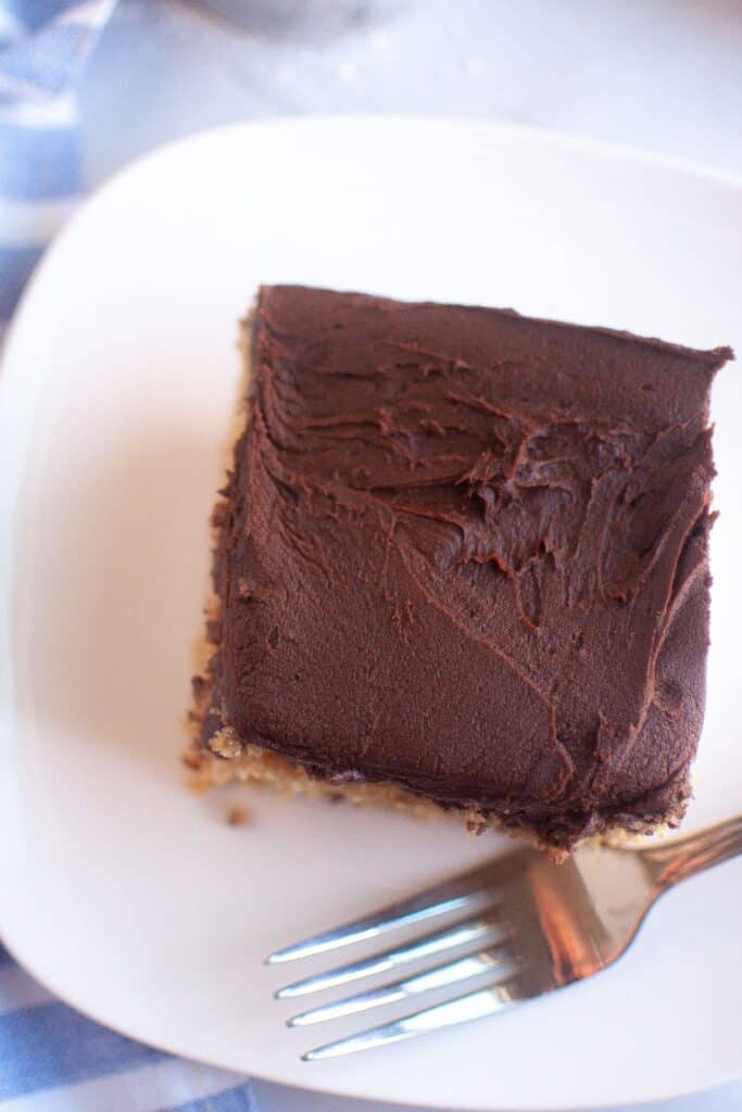 A close-up of a slice of Graham Cracker Cake topped with thick chocolate frosting on a white plate.