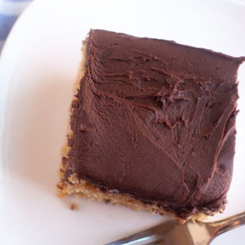 A square slice of graham cracker cake with a thick layer of chocolate frosting sits on a white plate, accompanied by a silver fork.