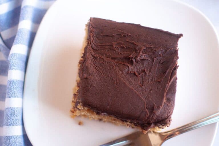 A square slice of graham cracker cake with a thick layer of chocolate frosting sits on a white plate, accompanied by a silver fork.
