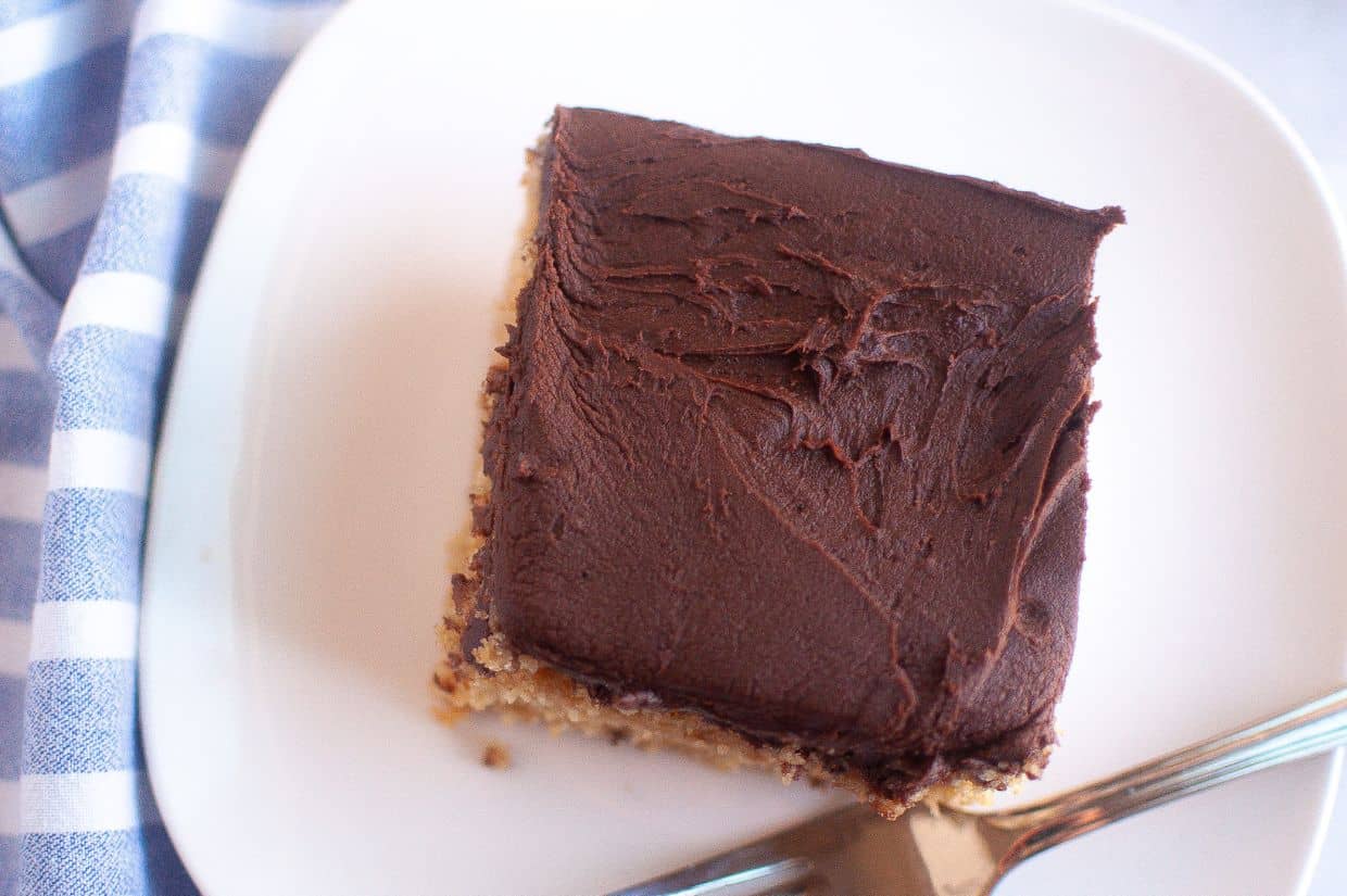 A square slice of graham cracker cake with a thick layer of chocolate frosting sits on a white plate, accompanied by a silver fork.