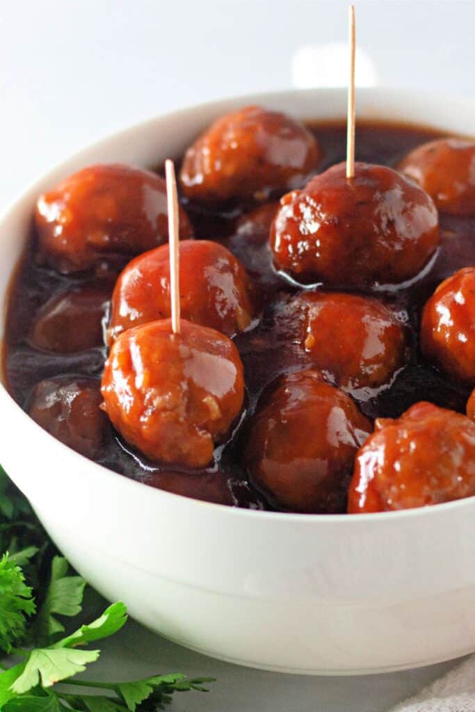 A white bowl filled with glazed honey garlic meatballs coated in a thick, glossy sauce. Some of the meatballs have toothpicks inserted, making them easy to serve. Fresh parsley is visible at the bottom left corner of the image.