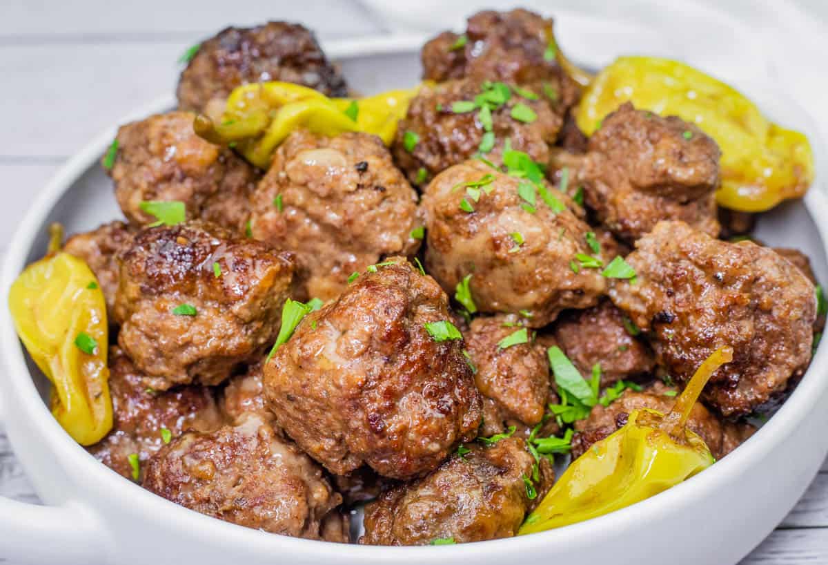 Bowl of browned Slow Cooker Mississippi Meatballs garnished with chopped parsley and yellow pepperoncini peppers.