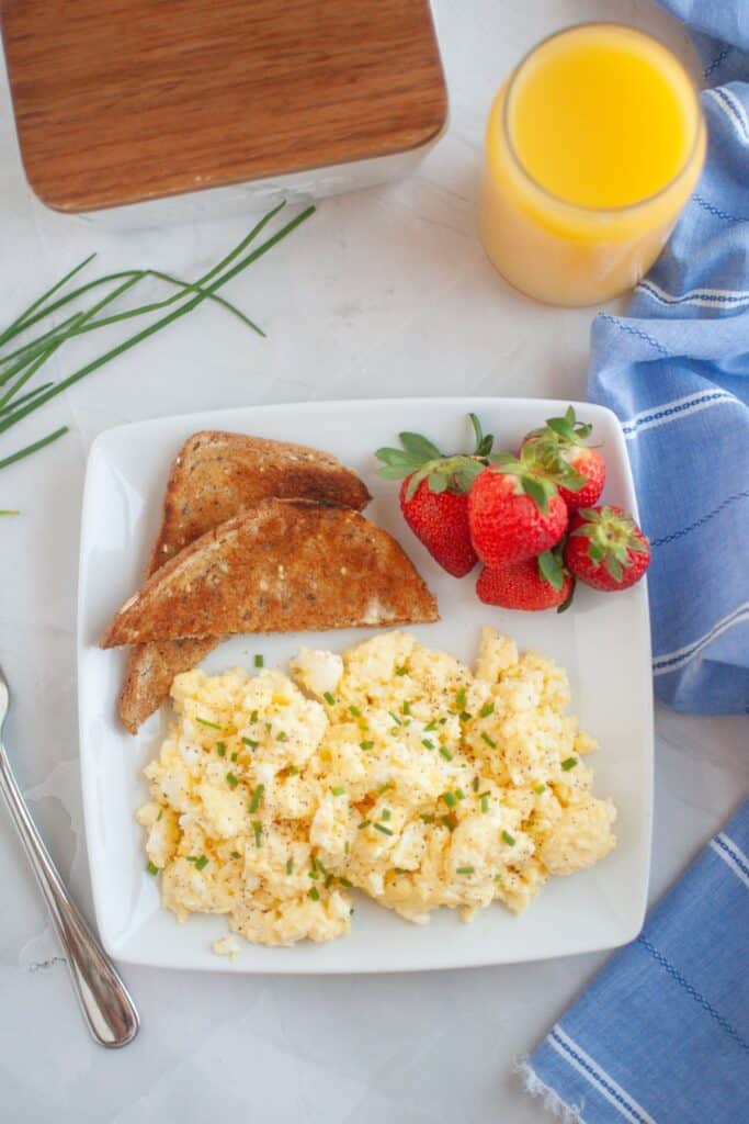This plate of scrambled eggs with cream cheese they are garnished with fresh herbs. The meal is completed with toasted whole-grain bread, fresh strawberries, and a glass of orange juice.