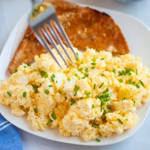 A plate of creamy scrambled eggs garnished with chopped chives, served with a slice of toasted whole-grain bread.