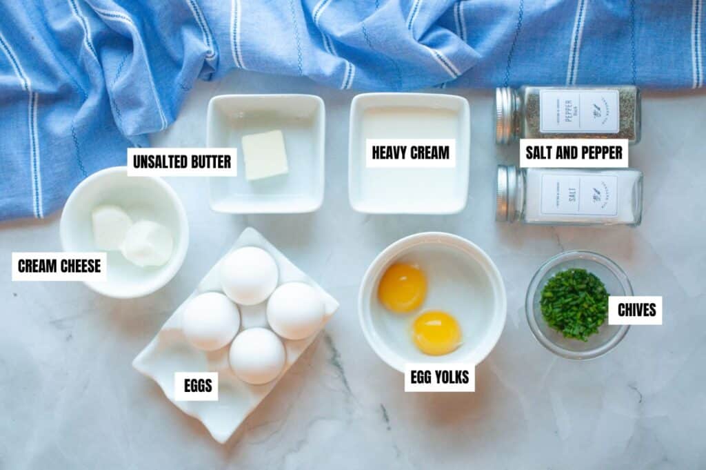 A flat-lay image of ingredients for scrambled eggs with cream cheese on a marble surface with a blue-striped cloth in the background.