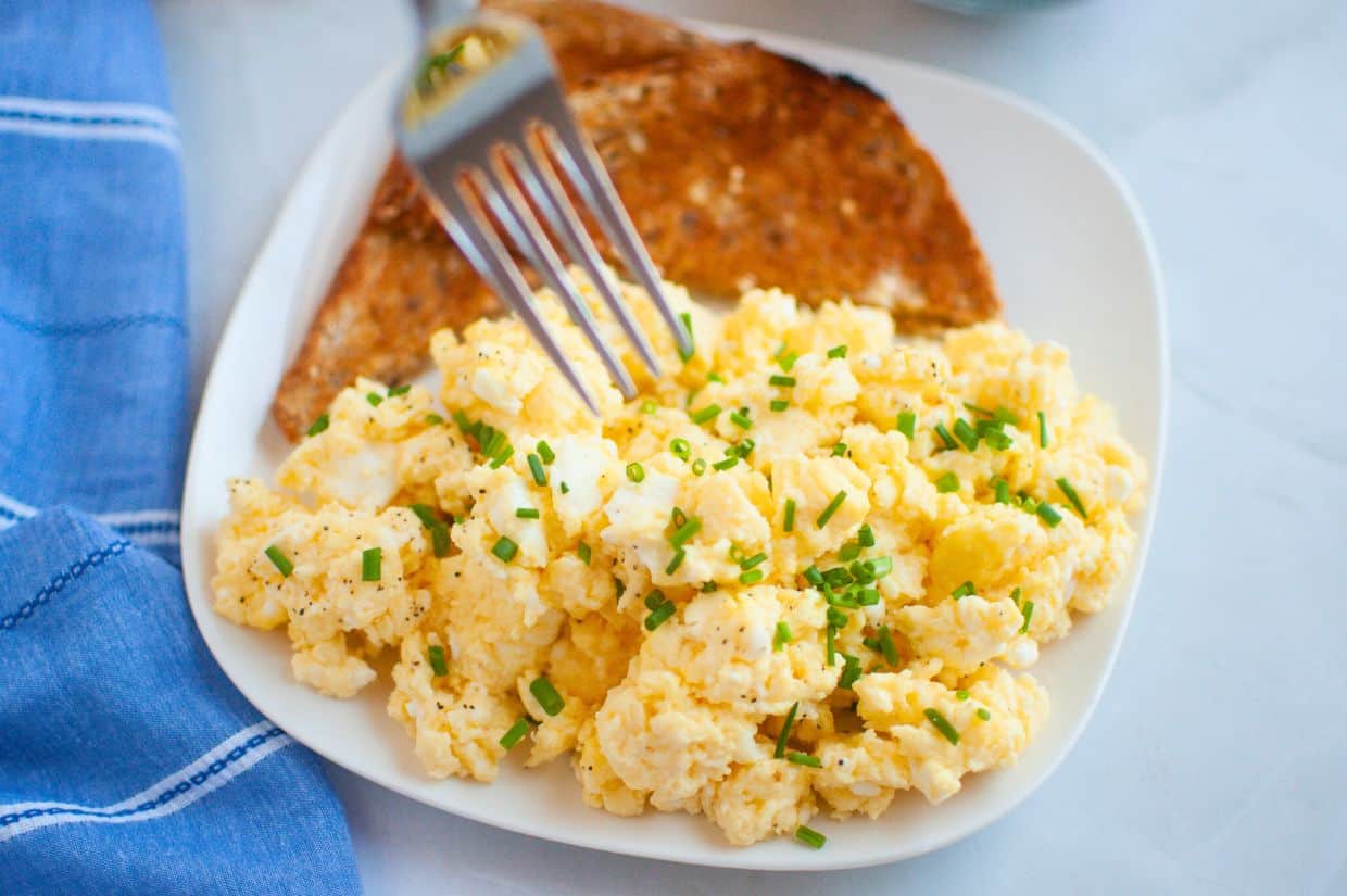 A plate of creamy scrambled eggs garnished with chopped chives, served with a slice of toasted whole-grain bread.