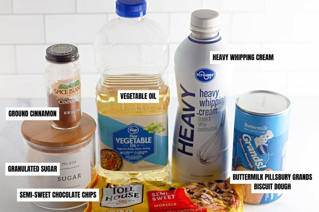 An assortment of ingredients for biscuit dough donut holes, including a can of Pillsbury Grands biscuit dough, a bottle of vegetable oil, heavy whipping cream, ground cinnamon, granulated sugar, and a bag of semi-sweet chocolate chips, all labeled and arranged on a white countertop.