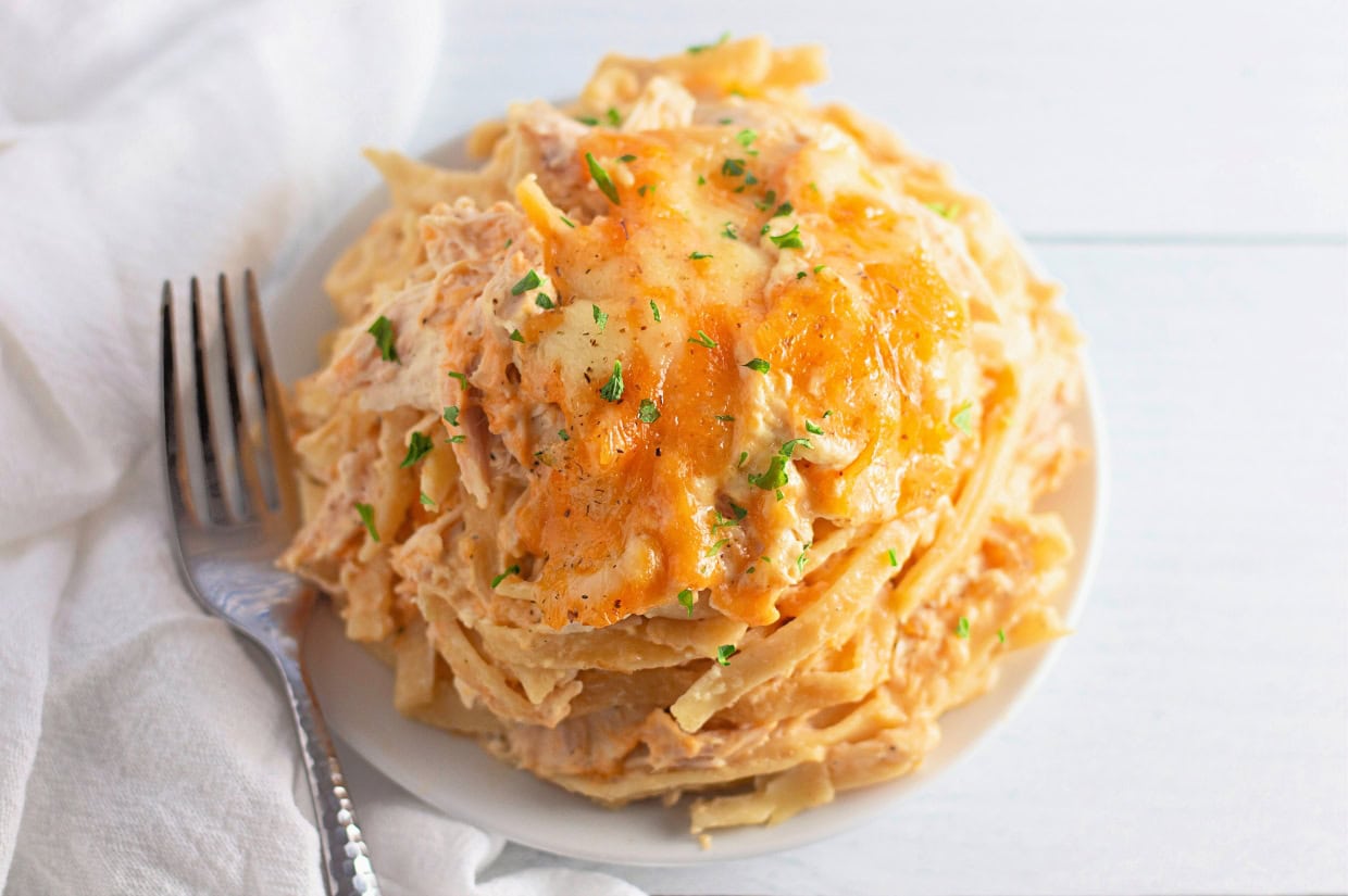 A hearty serving of creamy Chicken Tetrazzini is piled high on a white plate, topped with melted golden cheese and garnished with chopped parsley. A silver fork rests beside the dish on a white napkin, ready to dig into the rich pasta.