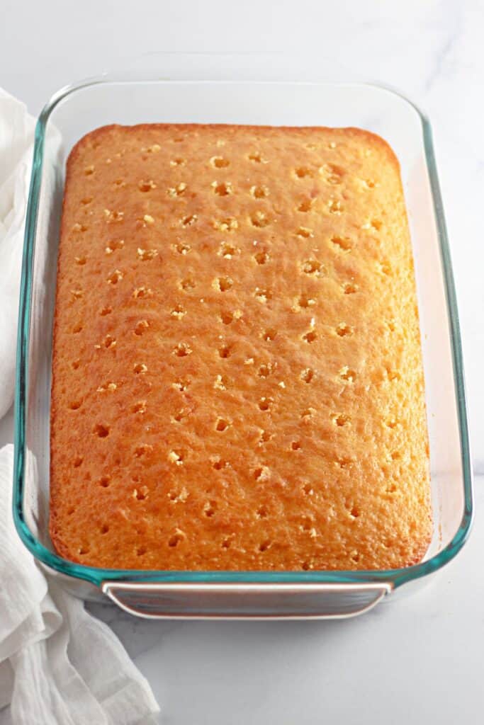 A freshly baked yellow cake in a glass baking dish with evenly spaced holes poked throughout, prepared for soaking in a sweet cinnamon-infused filling.