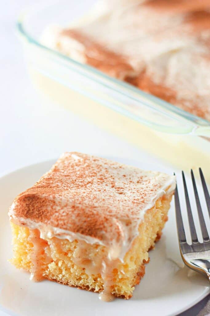 A slice of cinnamon roll poke cake on a white plate, topped with creamy frosting and a dusting of cinnamon, with sweet cinnamon filling oozing from the moist cake.