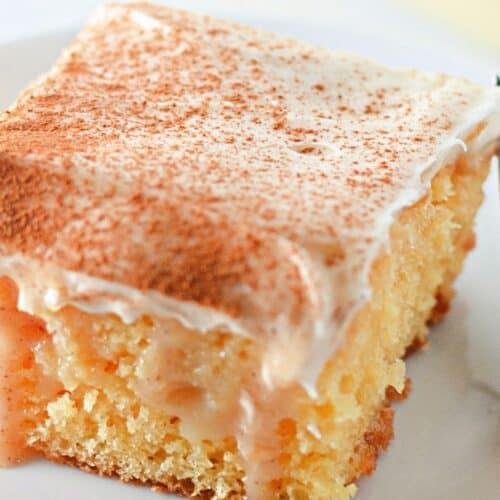 A close-up of a moist cinnamon roll poke cake with creamy frosting and a dusting of cinnamon on top, served on a white plate with a fork beside it.