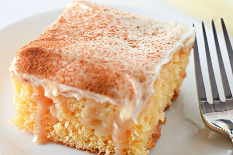A close-up of a moist cinnamon roll poke cake with creamy frosting and a dusting of cinnamon on top, served on a white plate with a fork beside it.