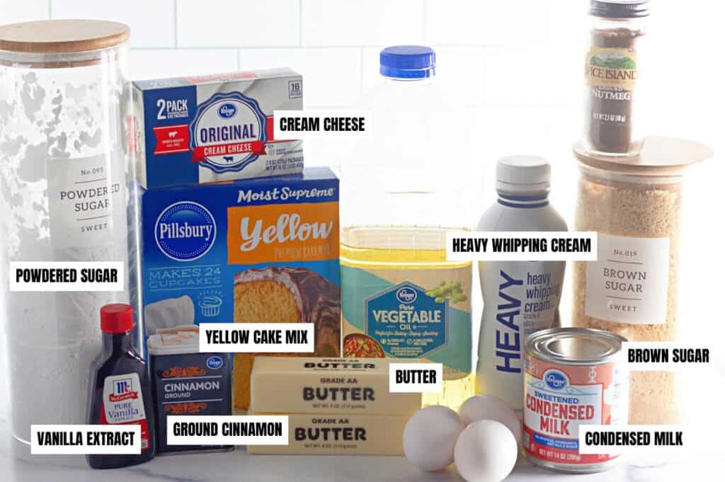 An assortment of labeled ingredients for a Cinnamon Roll Poke Cake, including yellow cake mix, cream cheese, condensed milk, heavy whipping cream, butter, eggs, brown sugar, powdered sugar, vanilla extract, ground cinnamon, nutmeg, and vegetable oil, arranged on a kitchen countertop.