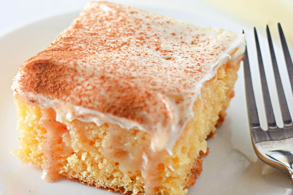 A close-up of a moist cinnamon roll poke cake with creamy frosting and a dusting of cinnamon on top, served on a white plate with a fork beside it.