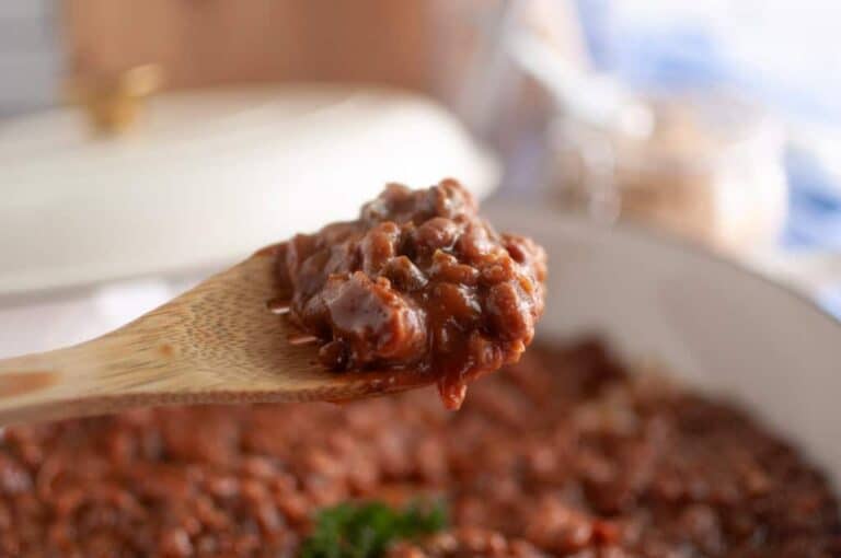 A wooden spoon holds a serving of Baked Bean Casserole above a pot filled with more bean casserole.
