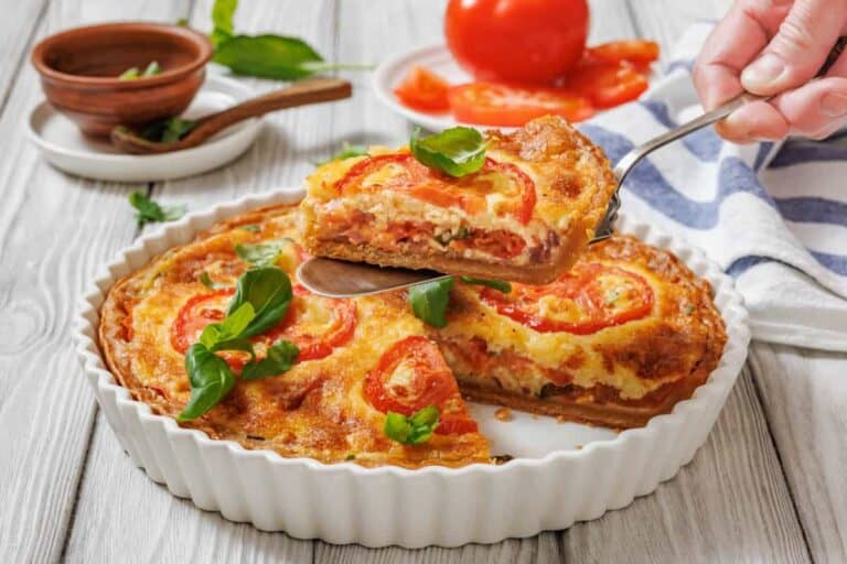 A slice of tomato pie being lifted with a serving utensil from a white fluted dish. A whole tomato pie is visible in the dish, garnished with tomato slices and basil.