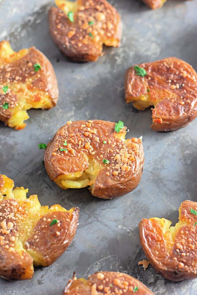 Crispy smashed potatoes with golden-brown edges, topped with Parmesan and fresh herbs, arranged on a baking sheet. The cracked surface reveals a soft, fluffy interior.