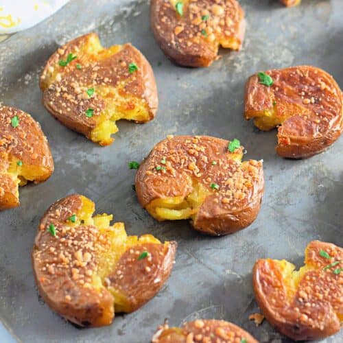 Golden-brown smashed potatoes with crispy edges, sprinkled with Parmesan and fresh herbs, rest on a baking sheet. The melted cheese and garlic seasoning add a rich, savory touch.