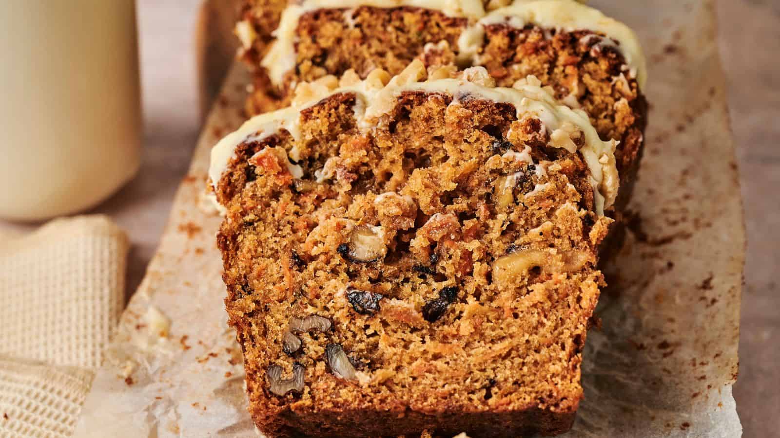 Close-up of sliced carrot cake loaf with visible nuts and raisins on a parchment paper, topped with a light icing drizzle.