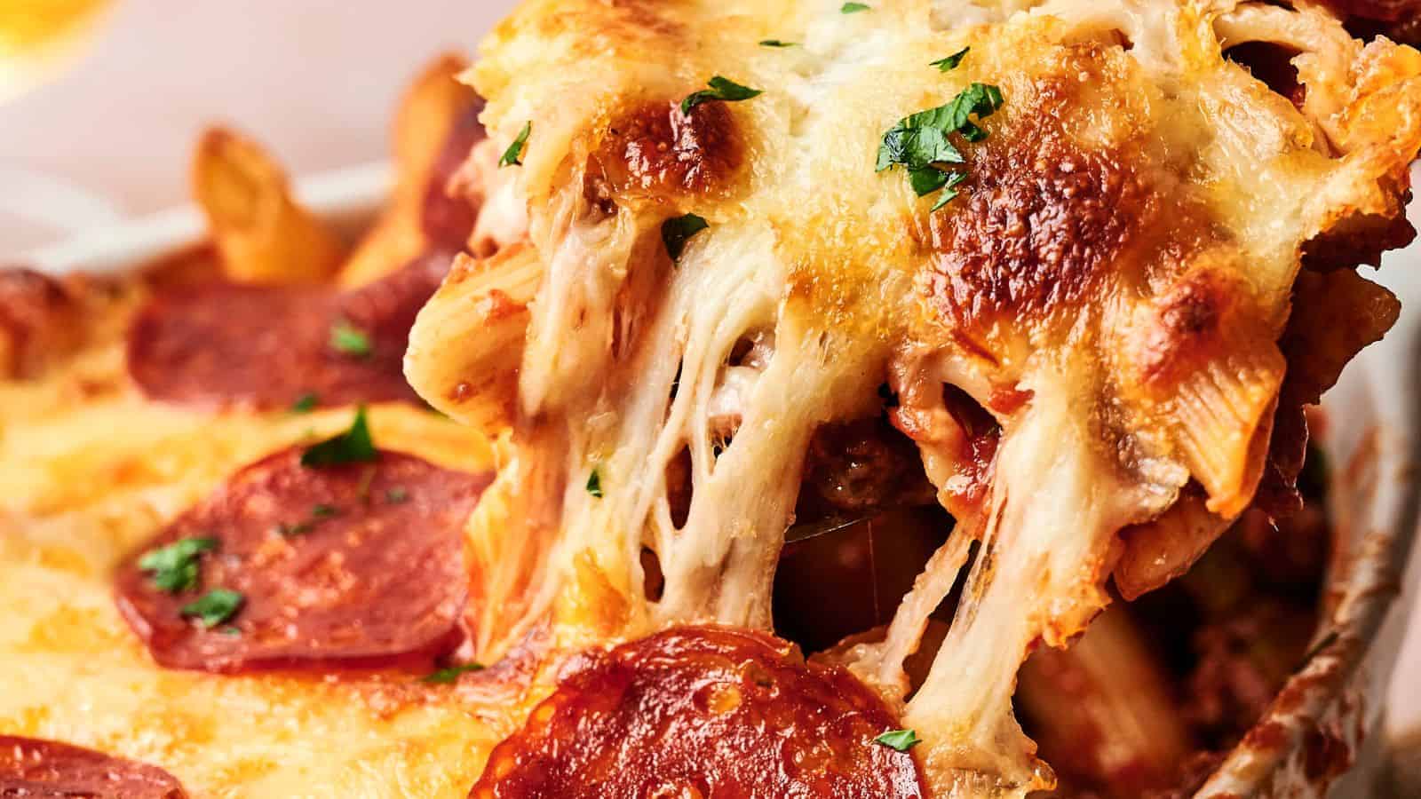 A close-up of a baked pasta dish with melted cheese, pepperoni slices, and herbs, showing a serving being lifted, revealing gooey cheese strands.