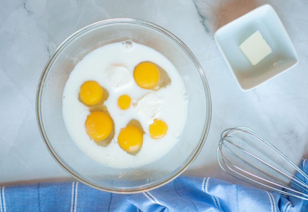 Glass bowl with egg yolks, cream cheese, and milk beside butter and whisk.