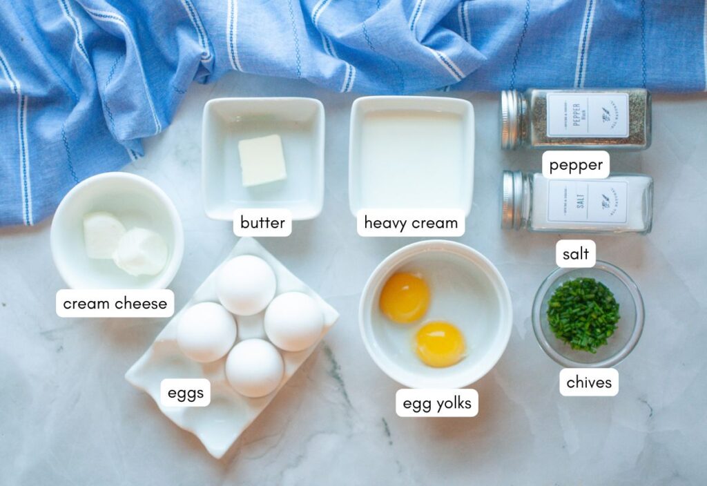 A flat-lay image of ingredients for scrambled eggs with cream cheese on a marble surface with a blue-striped cloth in the background.