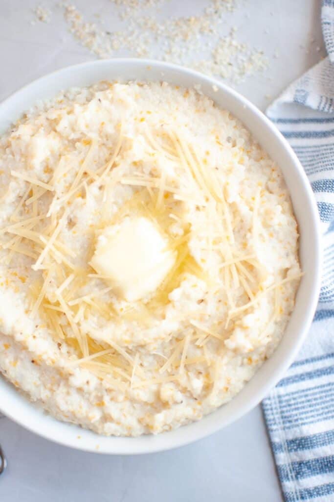 A bowl of creamy Southern-style corn grits topped with a melting pat of butter and shredded cheese, set on a light-colored surface with a striped kitchen towel beside it.