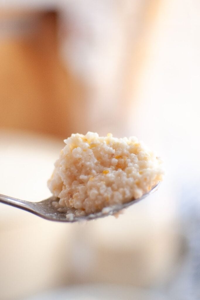 A close-up photograph of a spoonful of Southern-style corn grits, showing their creamy and slightly grainy texture with small yellow flecks of corn.