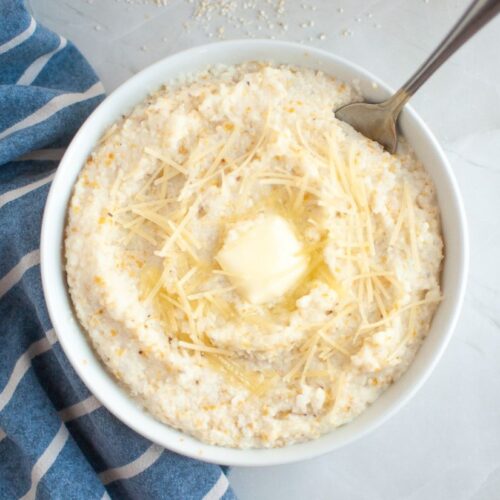 A bowl of creamy Southern-style grits topped with shredded cheese and a melting pat of butter, with a spoon resting inside the bowl.