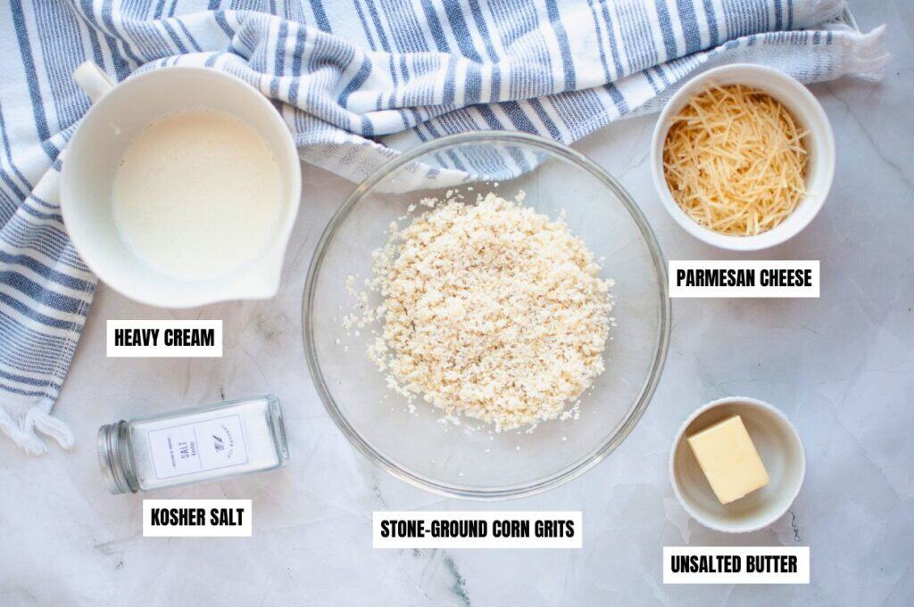A top-down view of ingredients for Southern-style corn grits, including stone-ground corn grits in a glass bowl, heavy cream in a small pitcher, shredded Parmesan cheese, a stick of unsalted butter, and a jar of kosher salt.