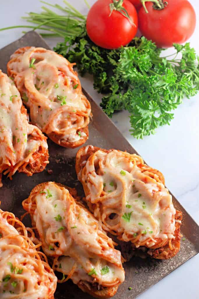 Slices of spaghetti bread topped with melted cheese and parsley, arranged on a baking sheet beside fresh tomatoes and parsley. A flavorful fusion of garlic bread and pasta, perfect for a hearty meal or snack.