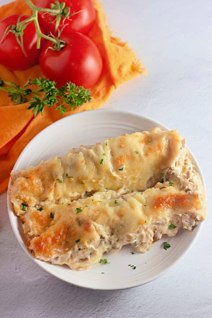 Two cheesy white chicken enchiladas sit on a white plate, their creamy filling peeking out from golden, bubbly edges. Fresh parsley adds a pop of green, with ripe tomatoes and an orange cloth in the background for a cozy, homestyle vibe.