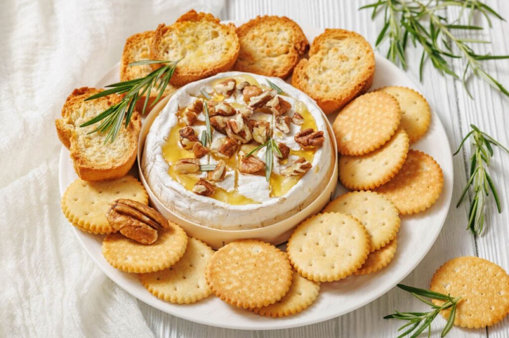 A round of baked brie topped with honey, walnuts, and rosemary is surrounded by golden crackers and toasted bread on a white plate, creating an inviting appetizer.