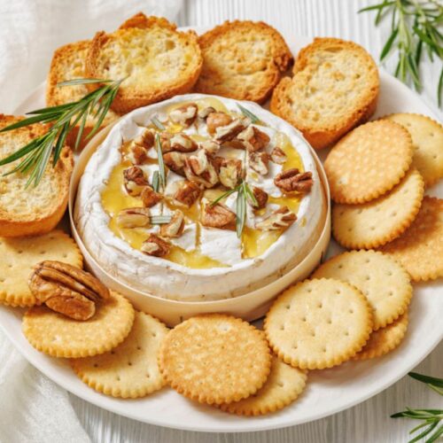 A round of baked brie topped with honey, walnuts, and rosemary is surrounded by golden crackers and toasted bread on a white plate, creating an inviting appetizer.