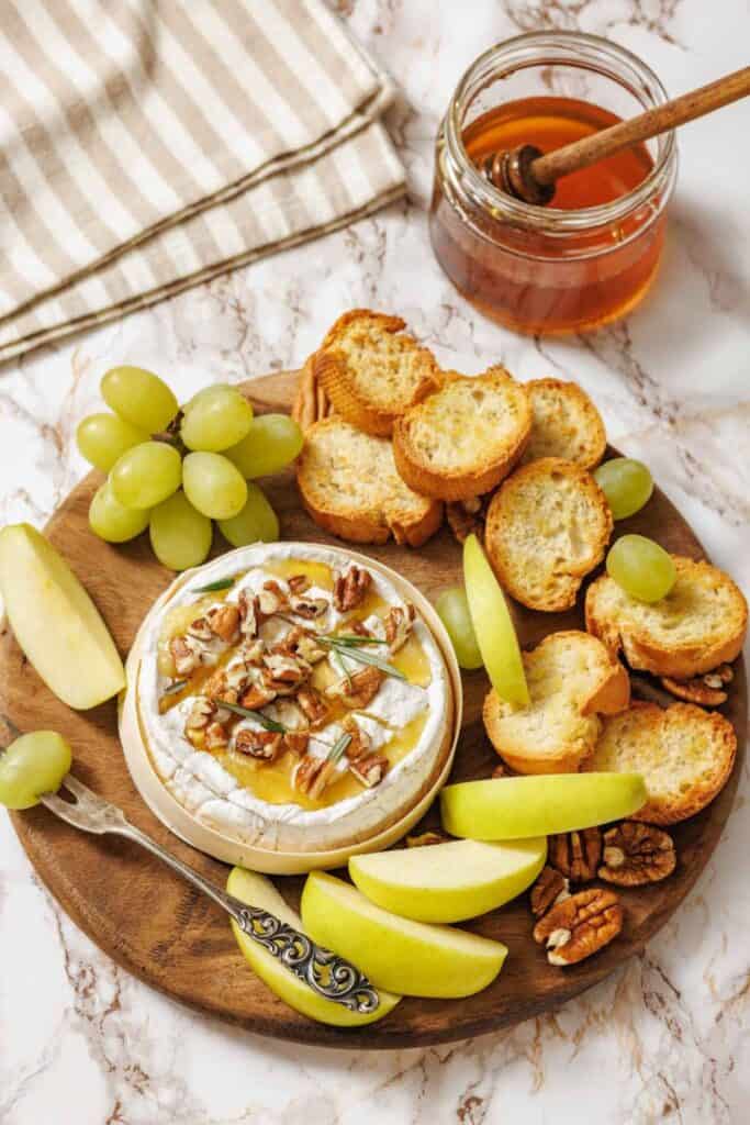 Round wooden platter with baked cheese topped with pecans and honey, surrounded by green apple slices, grapes, toasted baguette slices, and pecans, set on a marble surface with a jar of honey in the background.