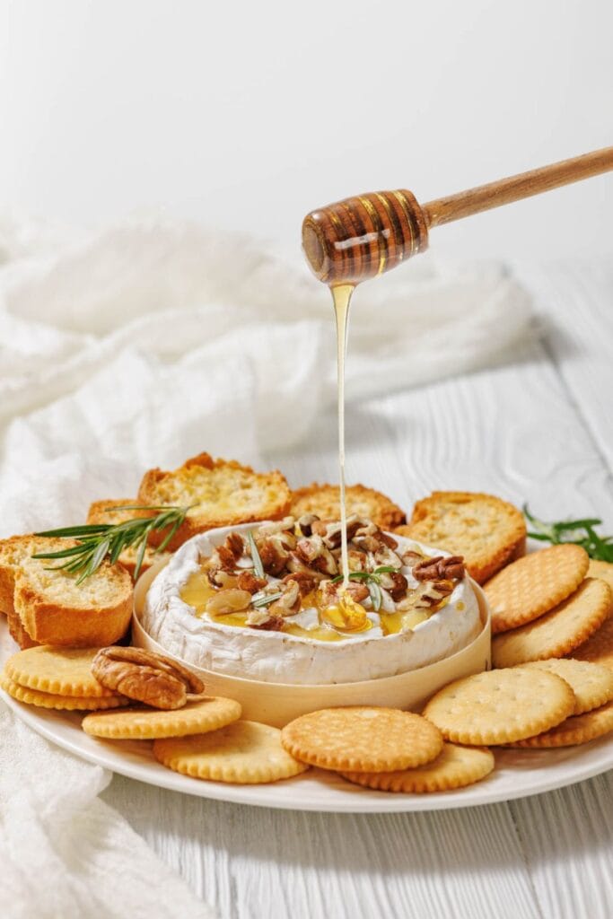 A cheese wheel topped with nuts and rosemary is drizzled with honey on a white plate surrounded by crackers and toasted bread slices. Rustic and appetizing.