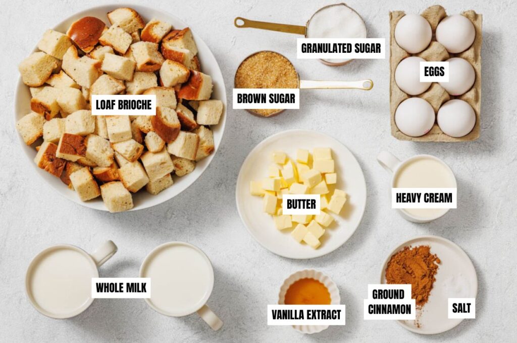 An array of bread pudding ingredients on a light surface: brioche cubes, sugars, eggs, butter, cream, milk, vanilla, cinnamon, and salt.
