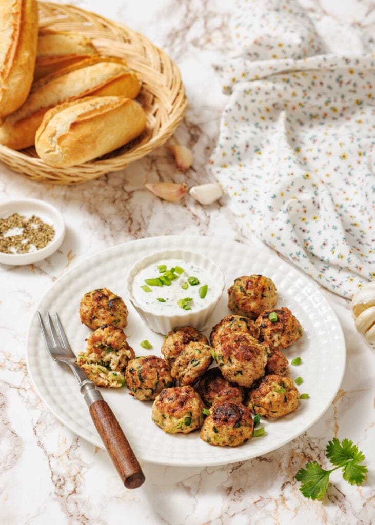 A plate of chicken zucchini poppers served with a small bowl of creamy dipping sauce topped with chopped green onions. A fork and a broken popper revealing the inside are on the plate, with a basket of baguettes, garlic cloves, and a floral napkin in the background.