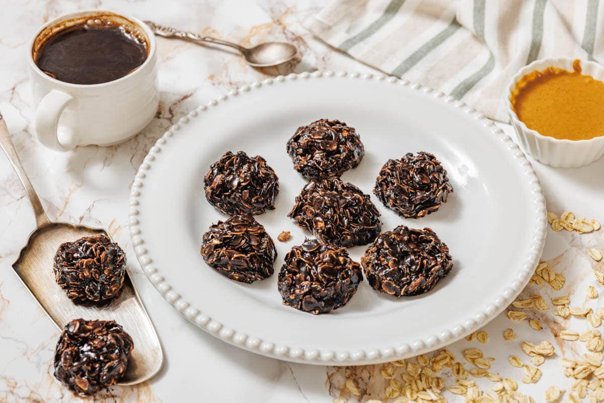 A white plate with seven Peanut Butter No-Bake Cookies on a marble surface, surrounded by a cup of coffee, a spoon, a bowl of nut butter, oats, and a striped cloth.