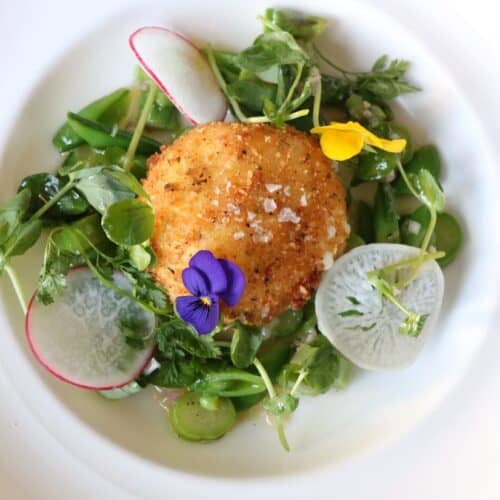 A golden, crispy fried burrata sits atop a bed of spring vegetables, including sliced radishes, snap peas, and leafy greens, garnished with edible flowers and drizzled with Meyer lemon vinaigrette.