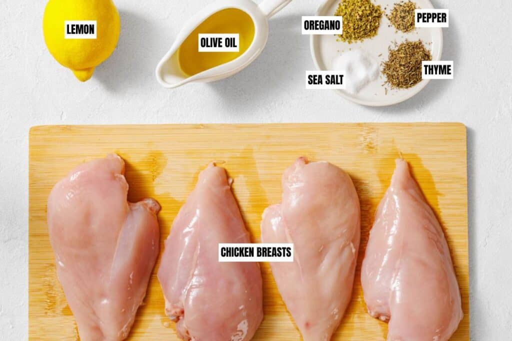 Raw chicken breasts on a cutting board surrounded by labeled ingredients: lemon, olive oil, oregano, thyme, pepper, and sea salt.