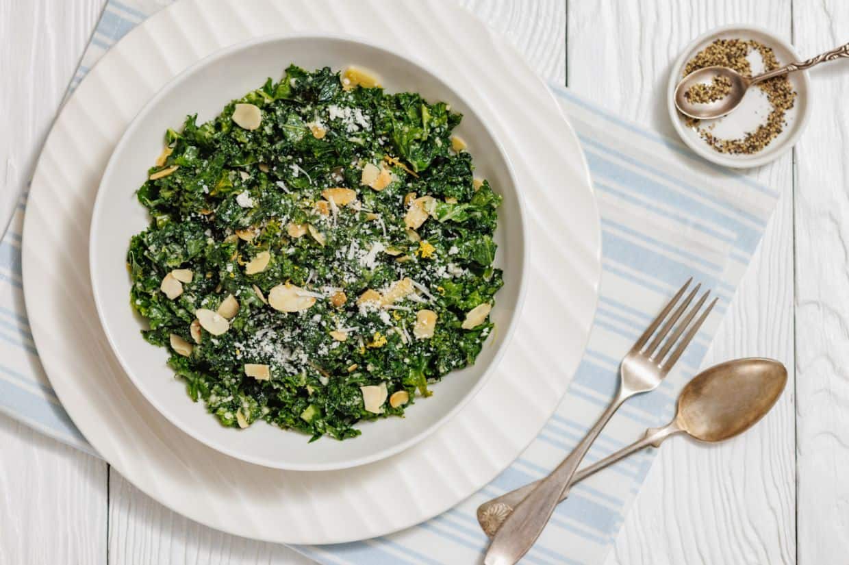 A bowl of fresh kale salad topped with lemon zest, shaved Parmesan cheese, and sliced almonds.