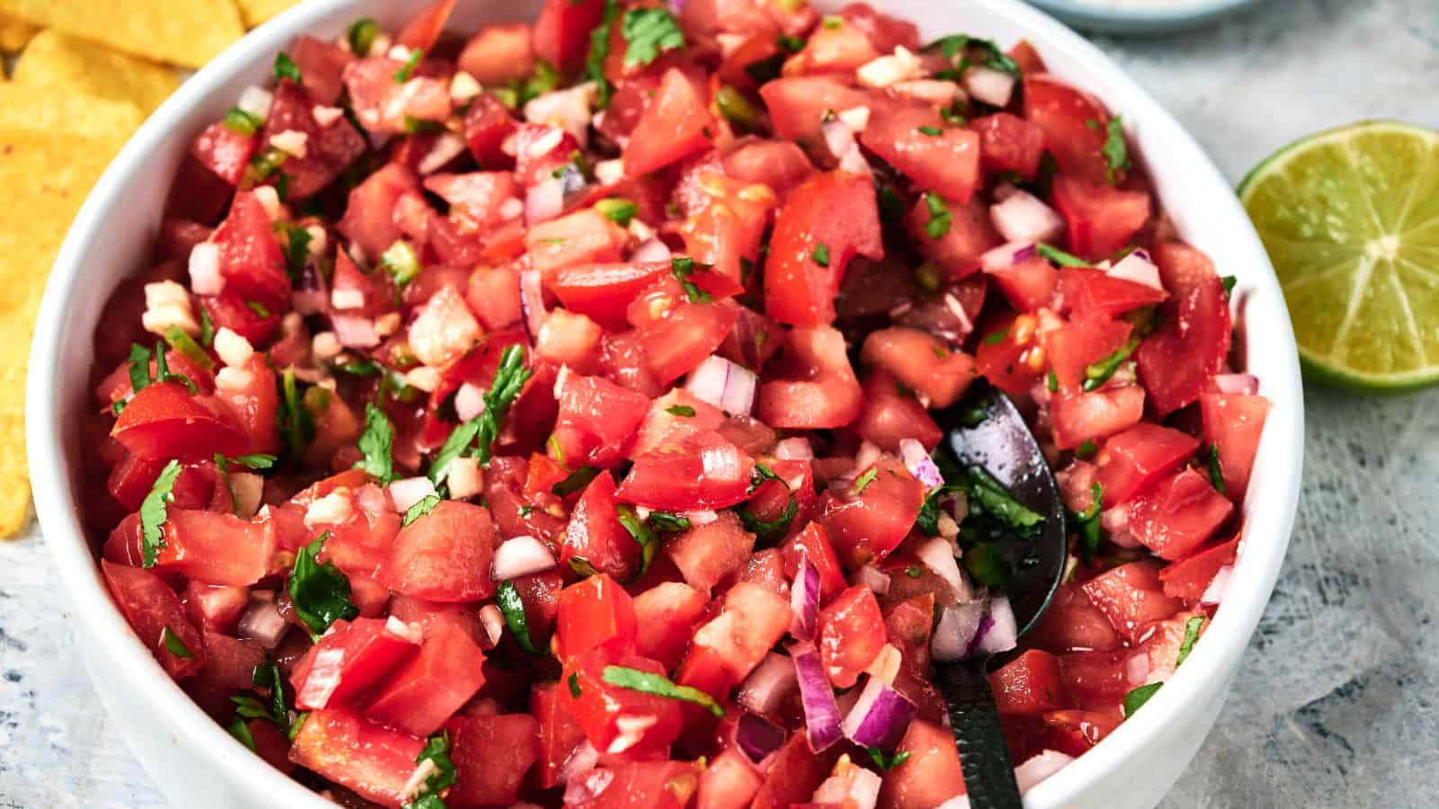 A bowl of pico de gallo with chopped tomatoes, onions, cilantro, and jalapeños with a spoon inside, next to a lime and tortilla chips.